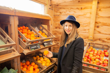 Woman in dark coat and navy hat holding an orange in her right hand at rustic farm shop with fresh fruits