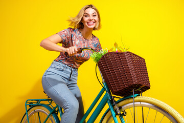Cheerful young woman enjoying a stylish ride on a colorful bicycle with flowers by a vibrant yellow...