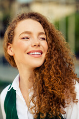 Confident young woman with red curly hair smiling outdoors on a sunny day surrounded by a cityscape
