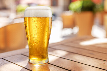 Glass of chilled beer on wooden table and blurred summer terrace at the background.