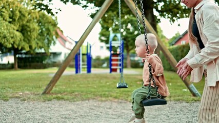 Mom on the playground swings her little son on a swing. Toddler sits on a swing and caring mother swings him. Mom and son walk in the fresh air. Happy family concept. Child development.