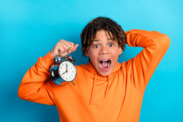Excited boy holding an alarm clock in orange hoodie on blue background, expressing surprise and...