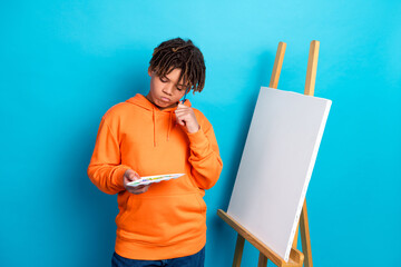 Young boy in orange hoodie holding a paint palette and standing by an empty canvas against blue...