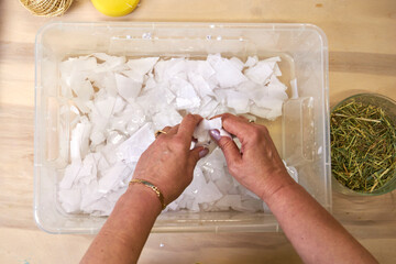 Woman placing torn white paper into water container for pulp making. DIY handmade paper preparation...