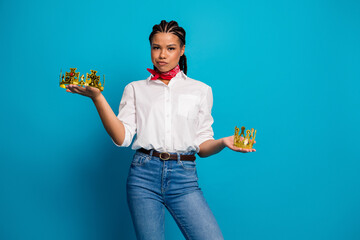 Stylish woman holding decorative crowns in hands against vibrant blue background demonstrating...