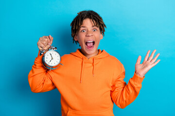 Excited boy in orange hoodie holding alarm clock with surprised expression against blue background