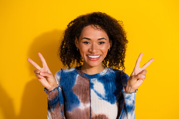 Happy young woman posing with peace signs on vibrant yellow background in casual outfit showcasing...