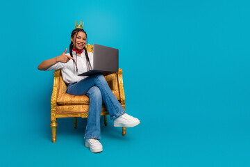 Young stylish woman with braids and scarf holding a laptop, sitting on an elegant chair against a...