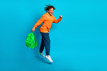 Joyful young boy jumping with a green backpack, wearing an orange hoodie against a vibrant blue...