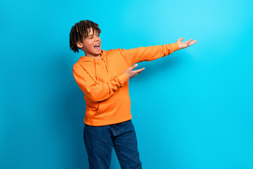 Cheerful boy in a bright orange hoodie gesturing against a vibrant blue background, expressing...