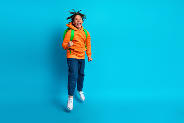 Excited young boy with backpack jumping on a vibrant blue background wearing colorful casual...