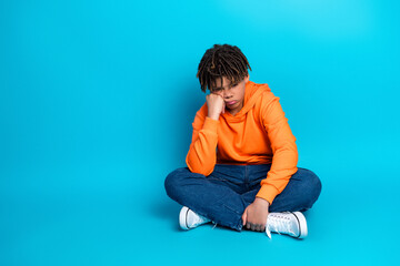 Teenage boy in casual orange hoodie sitting cross-legged on light blue backdrop, expressing...