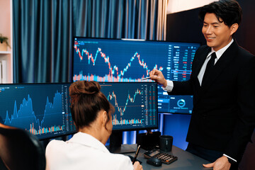 Smiling Asian business traders pointing to dynamic stock exchange data screen profit value online...