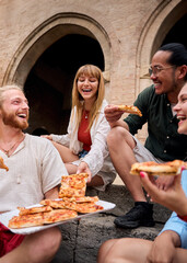 Vertical. Cheerful young friends enjoying delicious pizza together, laughing and sharing joyful moments while sitting on stone steps in an urban setting filled with vibrant city life