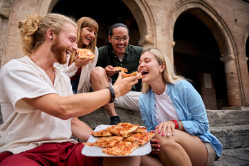 Cheerful young multi-ethnic friends laughing and sharing delicious pizza while sitting on steps in front of a stunning historical building in italy, enjoying a carefree moment together