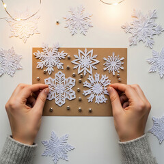 Top-down flatlay of hands arranging delicate white paper snowflakes and silver beads on kraft paper card. Warm ambient lighting, festive mood copy