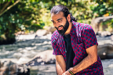 Obraz premium Man in plaid shirt with tied hair standing near river and extending arm toward colorful fence in lush Colombian forest at countryside hotel in Santuario, Risaralda, with space for text