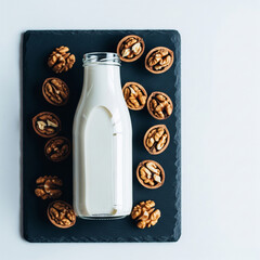Top-down minimalist shot of a glass bottle of walnut milk surrounded by scattered walnut halves on dark slate surface. Cool-toned light and space for text copy