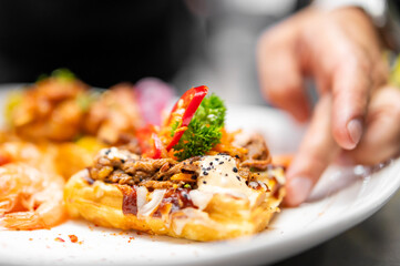 Gourmet waffle topped with pulled meat, shrimp, sauce, red peppers, and parsley, presented on a plate with a hand reaching toward the dish.
