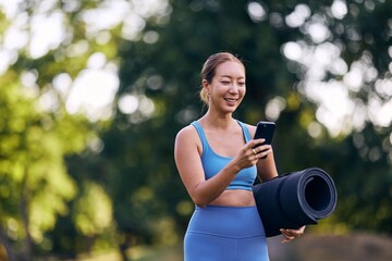 Woman Outdoors Holding Yoga Mat and Using Phone Amidst Lush Green Background