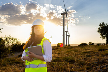 Female engineer holding tablet at wind farm during sunset with turbines in the background
