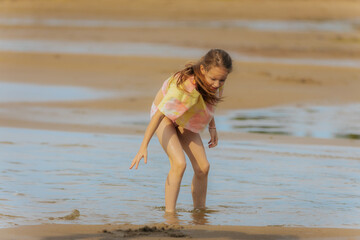 Fototapeta premium A young girl joyfully leaps into a shallow tidal pool on a beach, wearing a colorful shirt and shorts on a warm, hazy day.