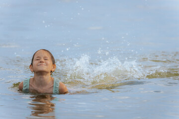 A happy young girl emerges from the water with her eyes shut and a wide smile, causing a big splash...