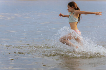 A joyful young girl in a swimsuit runs through shallow water at the beach, creating a large splash...