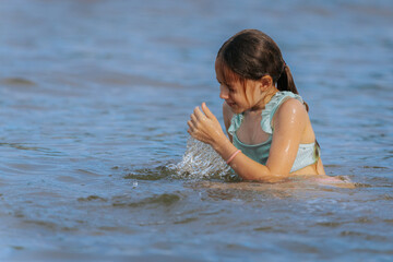 Fototapeta premium A smiling girl in a swimsuit splashes water playfully while sitting in a shallow lake or sea on a sunny day, enjoying summer fun.