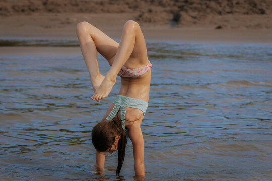 A young girl performs a handstand in shallow water at the beach, showcasing strength and playfulness during a sunny day. - Powered by Adobe