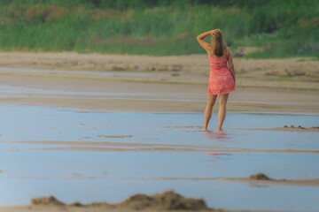 A woman in a red dress stands barefoot in shallow beach water, facing lush greenery in a peaceful...