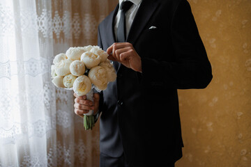 Groom holding a bouquet of white peonies in a formal setting before the wedding ceremony