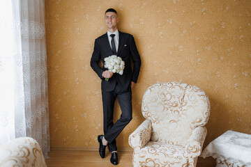 Groom poses with bouquet in elegant suit in cozy indoor setting before wedding ceremony