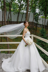 Bride in elegant white gown holds bouquet with flowing veil in scenic outdoor setting