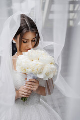 Bride holding bouquet of white peonies while wearing a stunning wedding dress in soft lighting