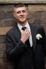 Groom adjusts tie while standing against stone wall at wedding reception venue