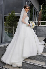 Bride in white gown carrying bouquet on steps outside a modern venue on wedding day