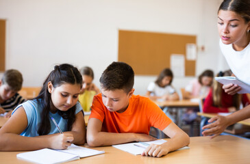Positive boys and girls studying and woman teacher helping in classroom
