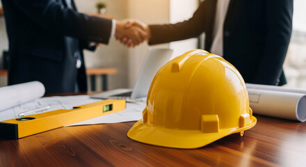 Yellow hard hat on wooden table, blurred background shows handshake, blueprints, symbolizing construction, partnership, agreement
