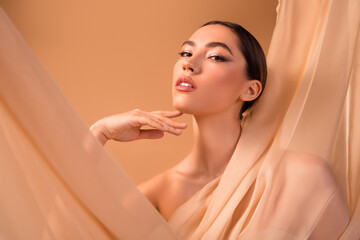 Beautiful woman with elegant makeup posing gracefully with beige fabric backdrop, emphasizing style...