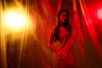 Glamorous young woman in red lighting with flowing dress and artistic cellophane background
