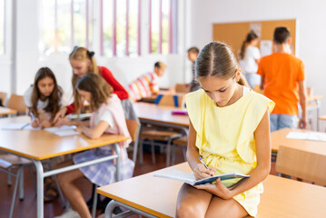 Portrait of young clever schoolgirl sitting on desk in classroom and looking at copybook in her...