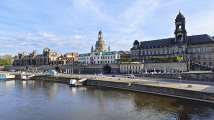 The river flows through the city in concrete banks. The embankment has piers and a roadway. Cars are driving. There are historic buildings and temples nearby. Sunny spring weather and blue sky