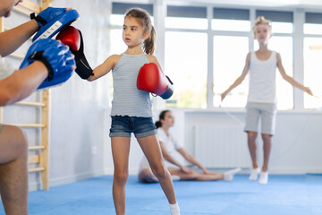 Concentrated sporty tween girl in boxing gloves practicing self-defense techniques with family in...
