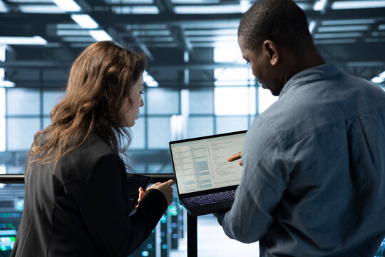 Programmers in data center workspace coding on laptop, analyzing metrics, optimizing storage volumes. Teamworking IT professionals in server hub using notebook, chatting, ensuring seamless operations