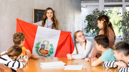 Decent teacher showing Peru flag to group of preteen schoolchildren in classroom during lesson