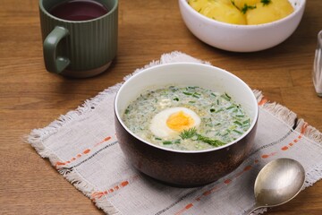 Cold sorrel soup with boiled egg and greens in a brown ceramic bowl on a wooden table. Served with boiled potatoes. Wild greens dishes.