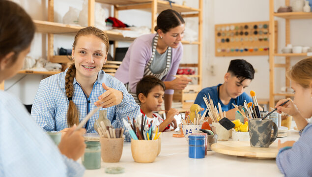 Joyful children, together with the teacher, paint ceramics with brushes at the table. Master class for children on pottery