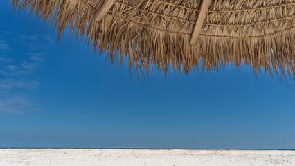 A fragment of a straw sun umbrella against a clear blue sky. Close-up. The upper part of the screen. The crossbars and the weave are visible. At the bottom of the frame is a white sandy beach.  