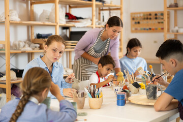 Children paint ceramics with brushes at the table in the creative studio. Teacher shows and helps children at the master class on ceramics